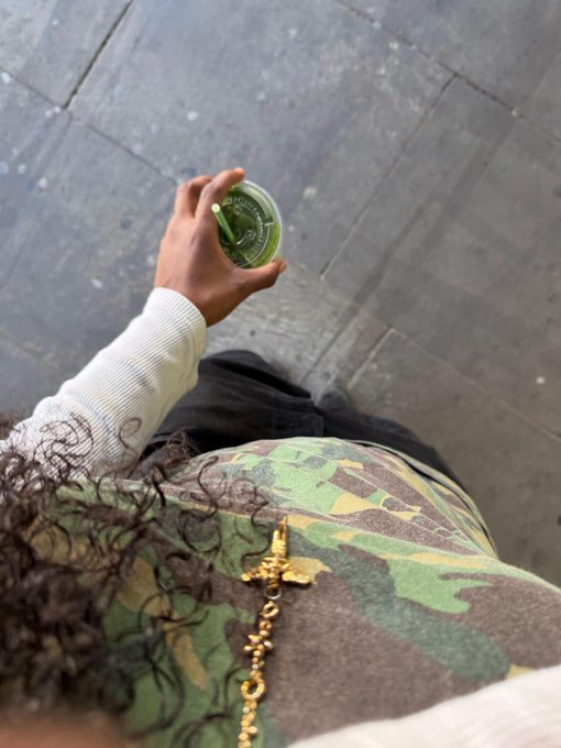 A person with curly hair wearing a camouflage top and gold bee pendant holds a green matcha or green tea beverage while standing on a stone sidewalk.