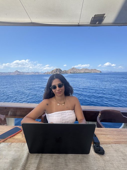 A woman wearing sunglasses and a white strapless top works on her MacBook at a wooden table on a boat, with a stunning ocean and island view behind her—capturing the perfect "remote office" vibe for the "GM frens, today's office" post.