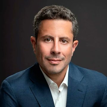 A professional headshot of Saifedean Ammous, a man with dark wavy hair wearing a navy blazer and white collared shirt, posed against a dark background. The portrait accompanies his provocative quote comparing Bitcoin's adoption to historical weaponized technologies like gunpowder rather than consumer innovations.
