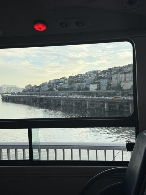 A view through a rain-spotted window of a vehicle, looking out over water toward a hillside city with buildings and vehicles on a bridge.