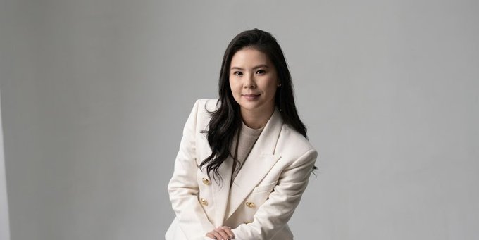 A portrait of an Asian woman with long dark hair wearing a cream-colored blazer with gold buttons over a beige top, posed against a gray background.