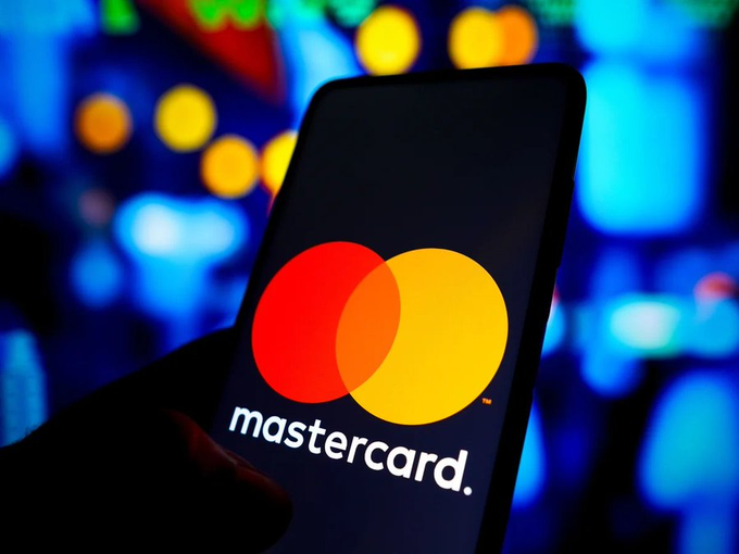 A smartphone displays the Mastercard logo (red and yellow interlocking circles) against a blurred background of blue and yellow lights.