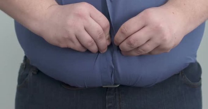 A close-up of a person wearing a blue button-up shirt straining over their stomach, attempting to button it over their belly, paired with blue jeans.