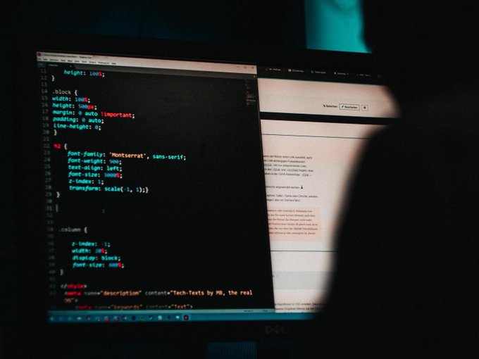 A dark photograph shows a computer monitor displaying CSS code (`.block`, `h1`, `.column` styling) on the left and a document on the right, partially obscured by a shadow.