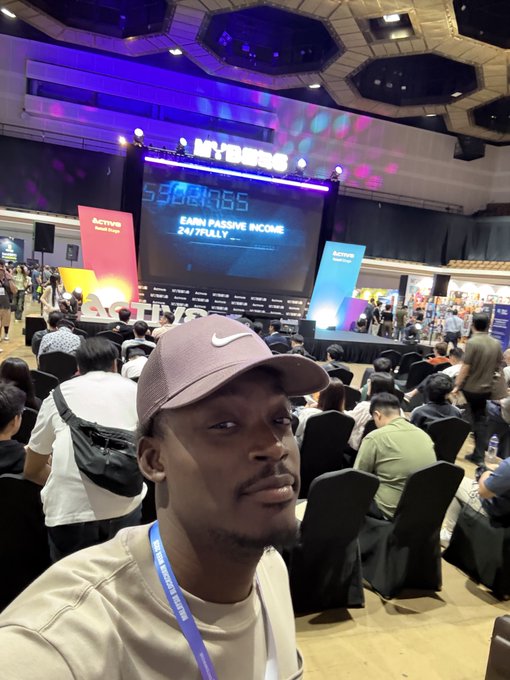 A selfie of a man wearing a Nike cap at what appears to be a Hybrid event, with a large screen displaying 'HYBRID EARN PASSIVE INCOME 24/7 FULLY...'.
