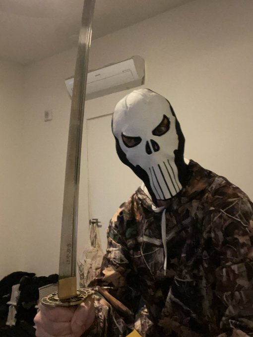 A person wearing a white skull mask and camouflage hoodie holds a large sword indoors.