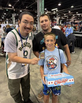A photo of two men and a young boy at what appears to be a convention, with the boy holding a 'Chicago' branded box of what looks like snacks or candy.