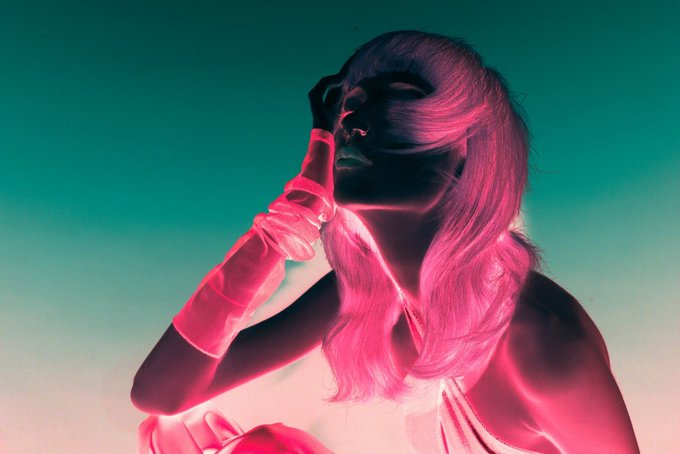 A stylized photograph of a woman with long hair bathed in vibrant pink and teal lighting, wearing long gloves and posing with her hand near her face.