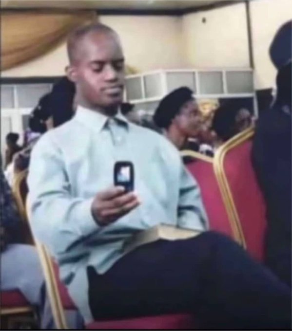 A young man in a light blue shirt sits on a red chair indoors, looking down at an early flip phone while holding a book, with a crowd of people seated behind him in what appears to be an event setting.