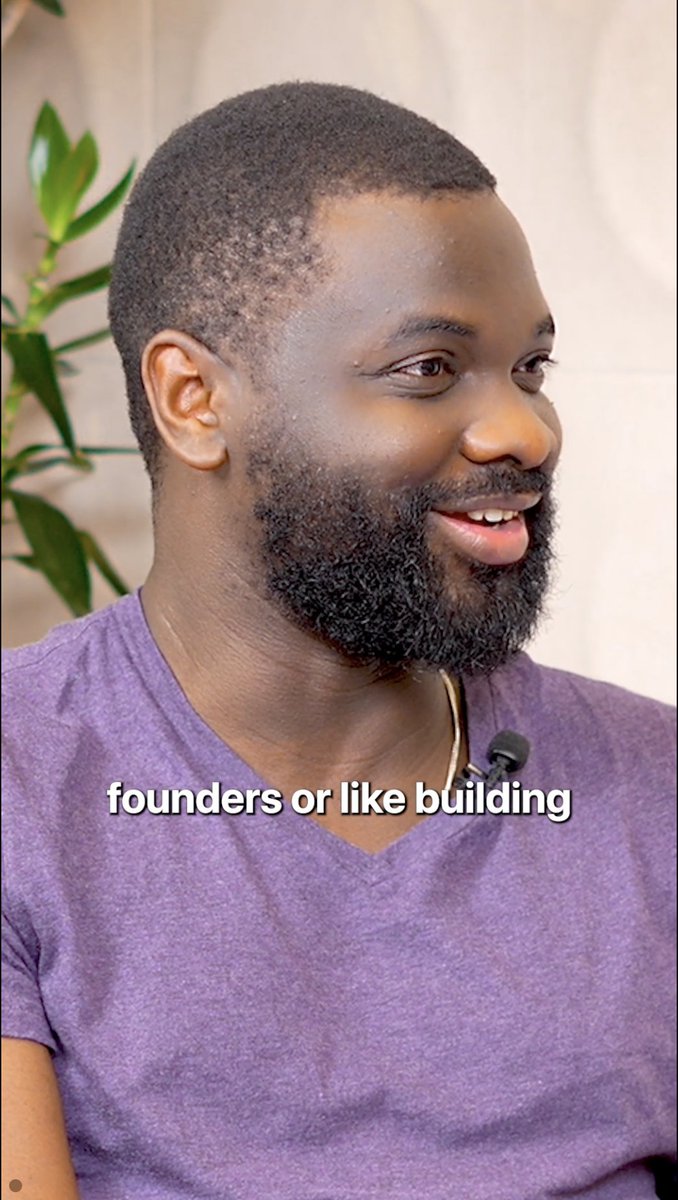 The image features a man wearing a purple shirt, smiling at the camera while sitting next to a plant. The background of the photo shows a wall that has text written on it, which reads " Founders or like building". This suggests that the man in the picture is likely discussing or promoting an idea related to building something, possibly a business or project.