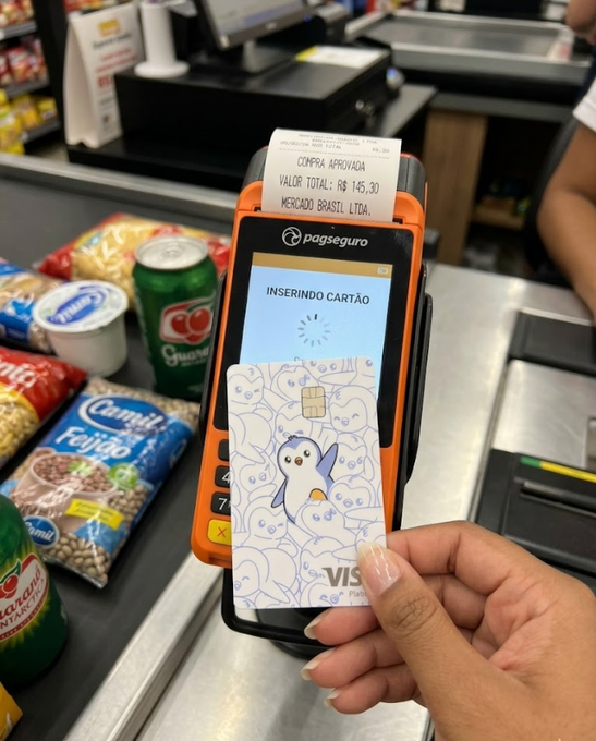 The image shows a person holding an orange wallet with a Visa card inside, which has a penguin design on it. The person's hand is visible above the wallet, while the wallet is placed below them. The background of the image features various items such as bags and food products like chips and candy bars. The post seems to be promoting stablecoins or cryptocurrency, suggesting that they are now part of everyday life for many people.