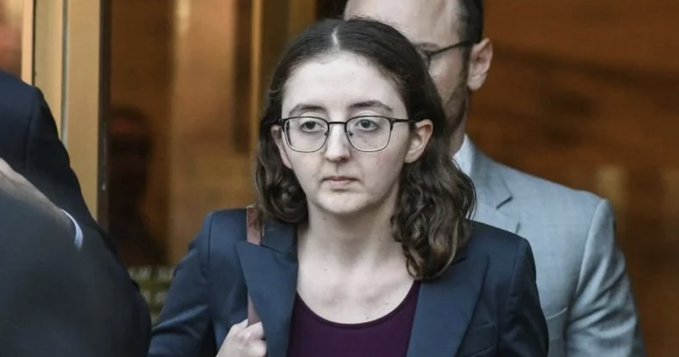 A young woman with glasses and dark hair, wearing a dark blazer over a maroon top, stands in what appears to be a formal setting with a man in a suit behind her.