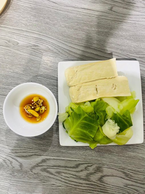An overhead photo of a meal on a wooden table featuring a white plate with tofu slices and bok choy, alongside a small bowl of soy-based dipping sauce with garlic.