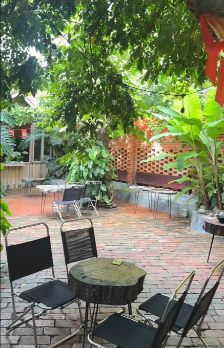 A serene outdoor café courtyard features a rustic wooden stump table surrounded by black metal chairs, shaded beneath lush green vines and tropical foliage, with a decorative red lattice wall in the background.

The image complements the post's message by providing a peaceful, plant-filled setting—likely a coffee spot ("ly cà phê")—where the poster can take a relaxing break before getting back to work on the X (Twitter) platform.