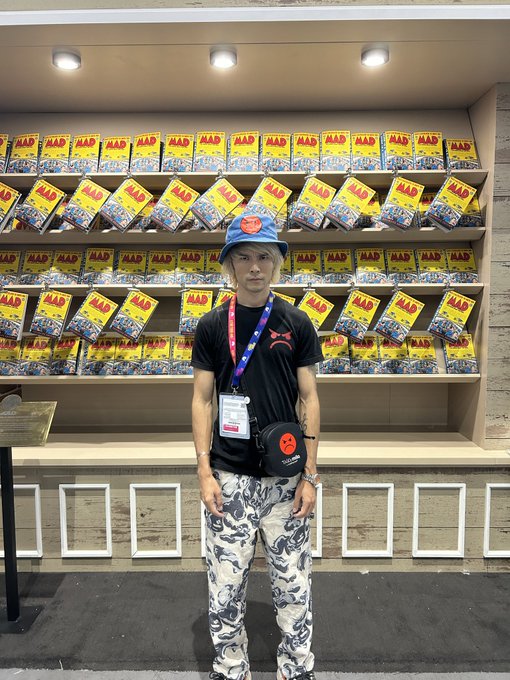 A young man wearing a blue bucket hat, a black Puma t-shirt, camouflage pants, and a small black crossbody bag stands in front of a display shelf stocked with yellow 'MAD' magazine issues.