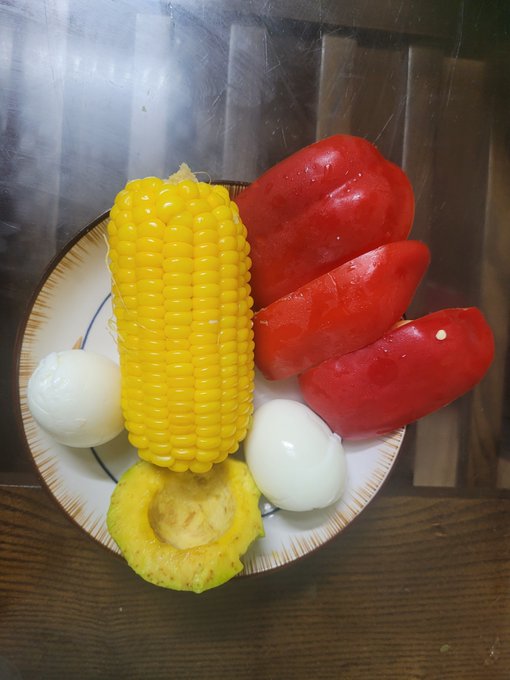 A plate containing a yellow corn cob, three red tomatoes, two boiled eggs, and a piece of squash.