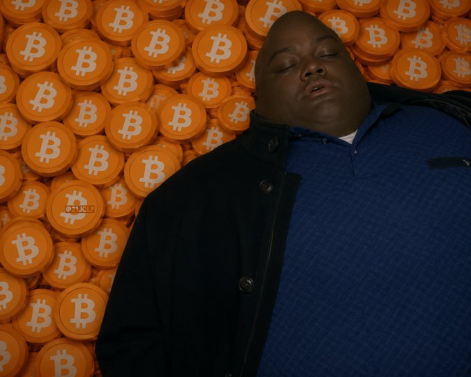 A person with their eyes closed leaning against a large pile of orange Bitcoin tokens, wearing a blue patterned polo shirt and dark jacket, with an NFL logo visible on one of the coins.