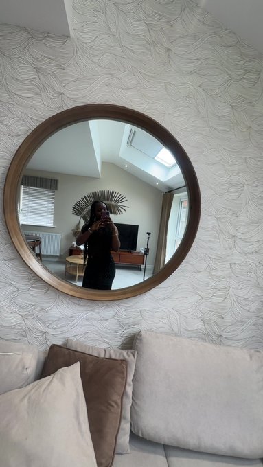 A woman takes a mirror selfie in a round, wooden-framed mirror mounted on a textured white wall, with a beige couch and pillows visible in the foreground.