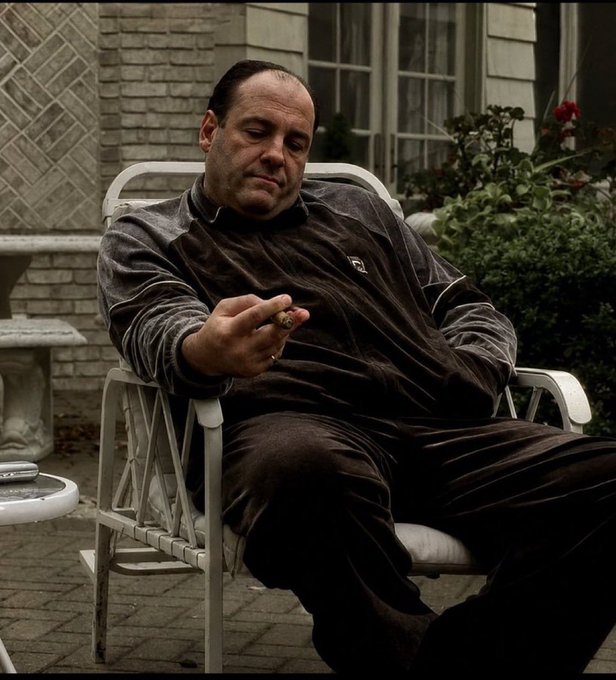 A man in a dark velvet tracksuit sits in a white metal patio chair, holding a cigar with his eyes closed, in a scene that appears to be from a television show.