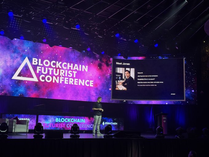 A presenter stands on stage at the "Blockchain Futurist Conference," with screens displaying the event logo alongside a speaker introduction titled "Meet Jonas." The scene captures a keynote presentation taking place on the Main Stage, featuring cosplay by Eterknow.
