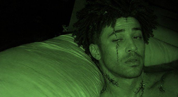 A moody, green-tinted scene shows a man with dreadlocks lying in bed, his face and neck marked with dark fluid trails.