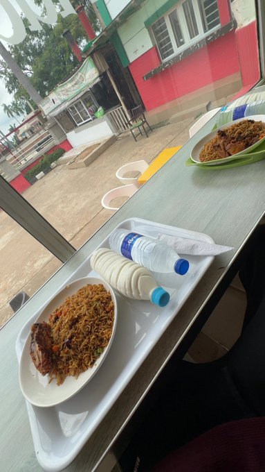 A tray of food with rice, fried noodles, a boiled egg, and bottled water sits on a window sill overlooking a red and white building.