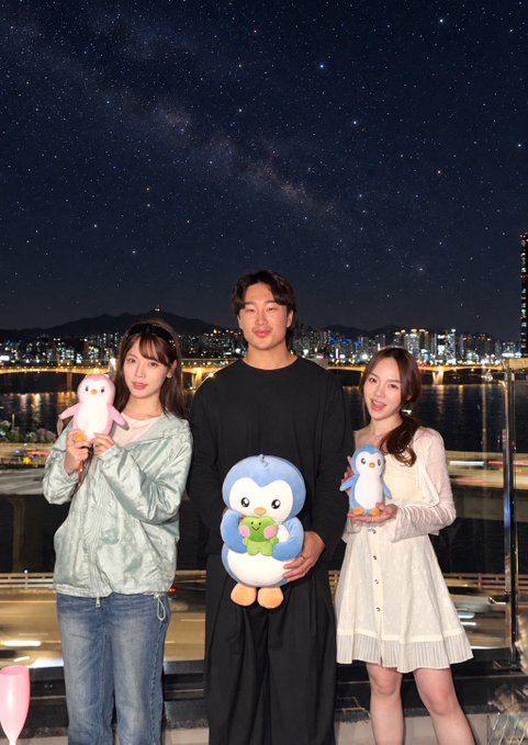 Three people pose on a rooftop at night holding plush toys—a penguin and a blue character with a frog—against a stunning backdrop of a starry sky with the Milky Way visible over a city skyline and water. The image captures the trendy, social atmosphere referenced in the caption celebrating Seoul's nightlife with the @fomo brand.