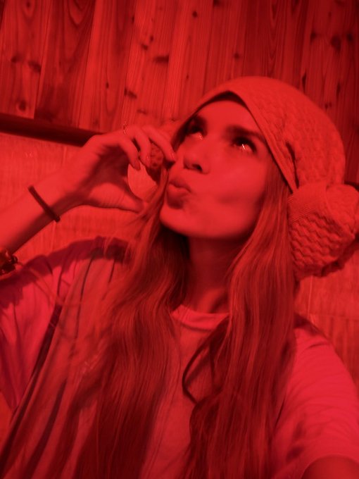 A selfie of a young woman with long hair wearing a knit beanie with a pom-pom, bathed in red lighting inside what appears to be a wooden sauna, kissing toward the camera.