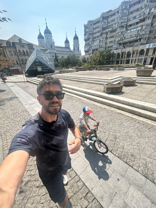 A man takes a selfie while a child rides a bike in a public square, with a large church visible in the background.