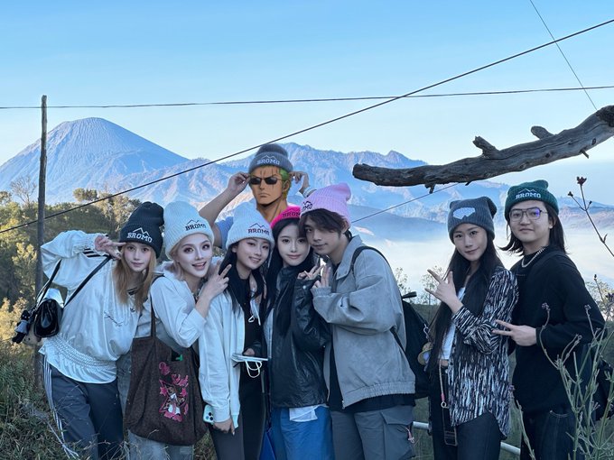A group of seven people wearing winter beanies and jackets posing for a photo with a snow-capped mountain (likely Mount Fuji) in the background.