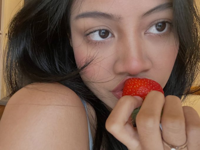 A close-up selfie of a dark-haired woman holding a fresh red strawberry near her mouth, looking directly at the camera.