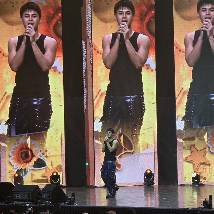 A male performer in a black tank top and studded shorts sings into a microphone on stage, while three large screens behind him display zoomed-in close-ups of the same performance. The image aligns with the post's reference to a "sold out" event, showcasing the singer's toned arms and confident stage presence amplified across the venue's screens.
