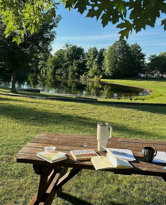 A wooden picnic table with an artist's setup—watercolor palette, sketchbook, mug, and thermos—sits on a grassy lawn beside a tranquil pond with ducks, framed by large trees.