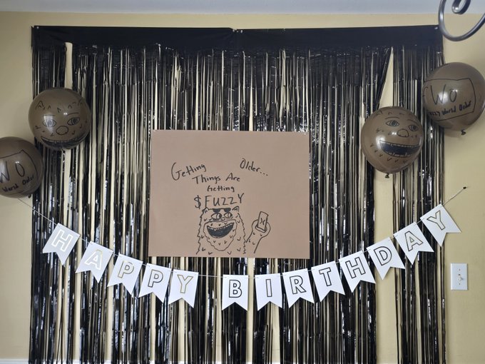 A birthday decoration setup features a hand-drawn sign reading "Getting Older... Things Are Getting FUZZY" with a cartoon furry creature, alongside a "HAPPY BIRTHDAY" banner and balloons marked "NO," playfully celebrating the man's milestone with crypto-themed humor that matches his "Fuzzy" and bearish puns.