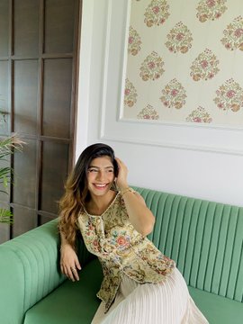 A smiling woman with long dark hair, wearing a floral top and white skirt, sits on a green ribbed sofa against a wall decorated with framed floral wallpaper.