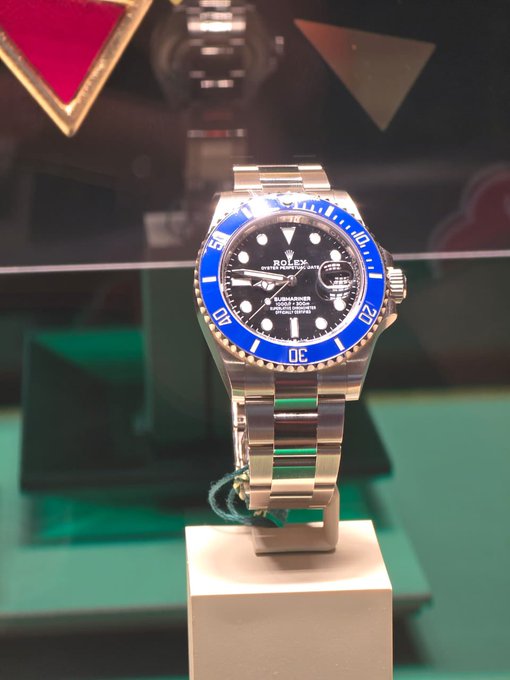 A Rolex Submariner watch with a distinctive blue bezel and black dial is displayed on a white stand.