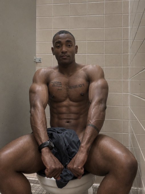 A shirtless, muscular man sitting on a toilet in a tiled bathroom, flexing his arms and looking directly at the camera, with chest tattoos visible.