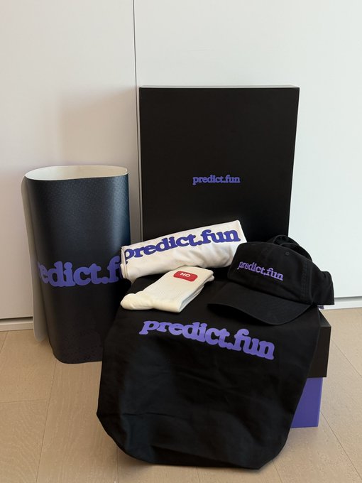 A black box with a purple lid and white label, containing a black t-shirt, white socks, and a black hat, is placed on a wooden floor against a white wall.