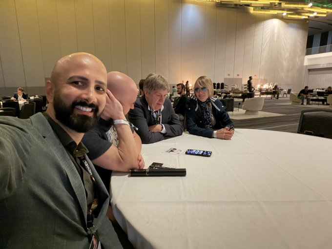 A group of people smiling for a selfie at a conference or event, seated around a round white table with a black Canon product box in the foreground, while other attendees can be seen mingling in a large convention hall background.
