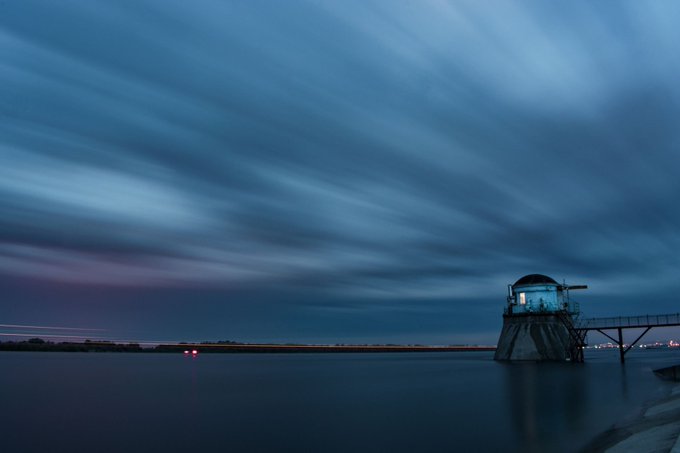 A long-exposure photograph of a twilight seascape with streaking clouds above calm water, featuring a small illuminated structure on stilts to the right.