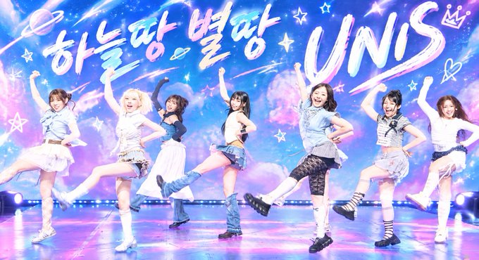 Seven female performers in coordinated white and blue outfits pose with raised arms on a colorful stage with Korean text and 'UNIS' written in the background.