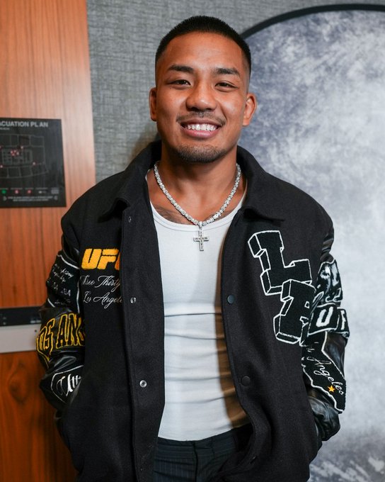 A young man with a short haircut smiles warmly at the camera, wearing a black varsity jacket featuring "UFC," "Los Angeles," and "LA" graphics over a white tank top, accessorized with a silver cross necklace. The image appears to be taken at the UFC Store, fitting the post's caption referencing a "Fitted for LA" promotion tied to Crypto.com and UFC 331.