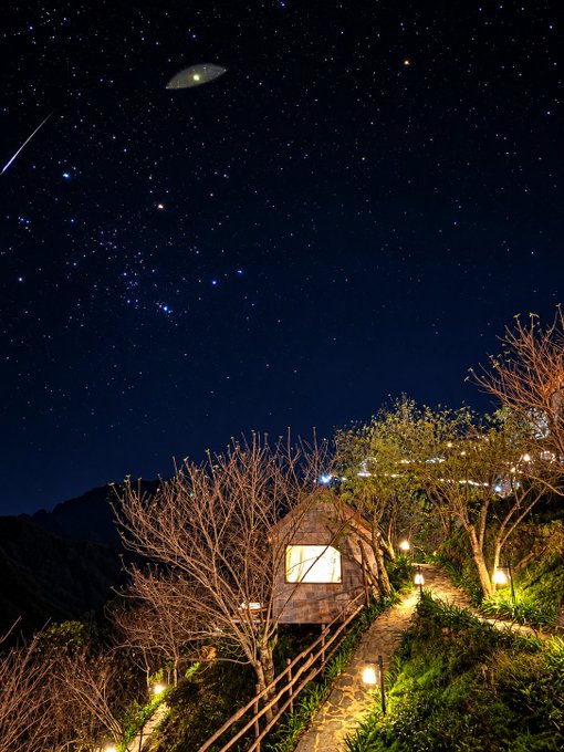 A solitary cabin glows warmly beneath a starry night sky featuring a UFO and shooting star, with a winding path illuminated by lantern lights. The image evokes the post's theme of solitary retreat and self-reliance, using the remote, peaceful setting to symbolize stepping away from the world to find comfort and clarity within oneself after emotionally taxing times.