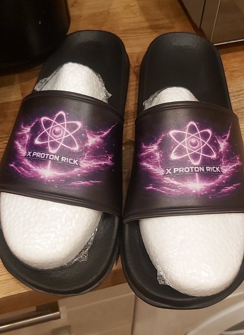 A pair of black slide sandals featuring a purple atomic design and the text "X PROTON RICK" sits on a wooden floor, humorously connecting the "XProtonRick" token from the post to wearable crypto-themed merchandise.