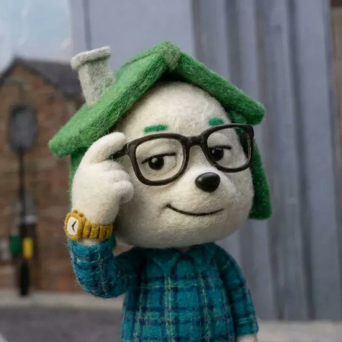 A felted or fabric stop-motion style figure of a character with green hair, large glasses, a blue plaid shirt, and a yellow wristband, adjusting their glasses in a thoughtful pose.