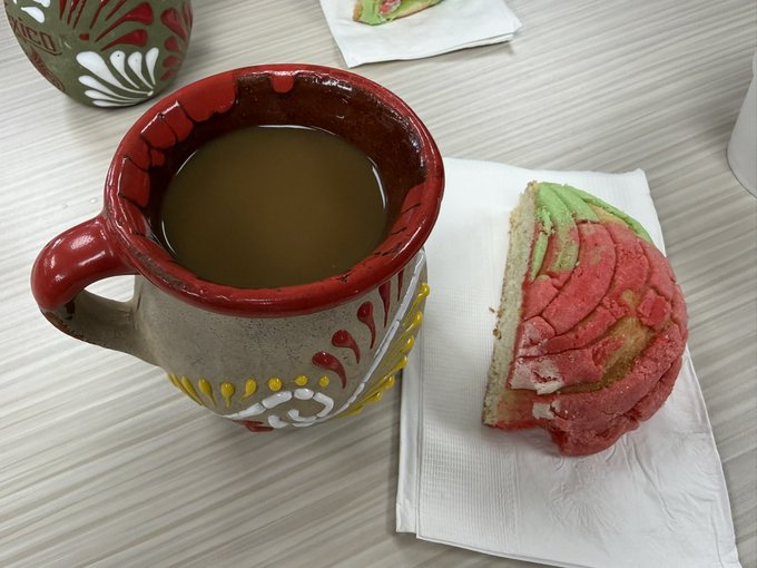 A photo of a rustic ceramic coffee mug filled with dark coffee, accompanied by a slice of colorful pink, green, and yellow concha bread on a napkin.