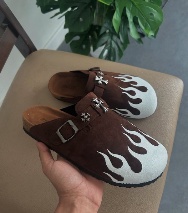 A hand holds a pair of brown suede clogs featuring white painted flame designs and decorative silver concho embellishments, with the brand name "GRACOSY" visible on the footbed.