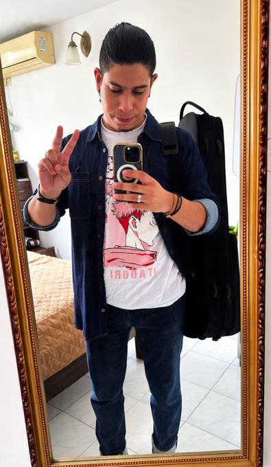 Mirror selfie of a young man in a denim shirt over a graphic tee, holding a peace sign and phone, wearing a backpack in a room with a bed.