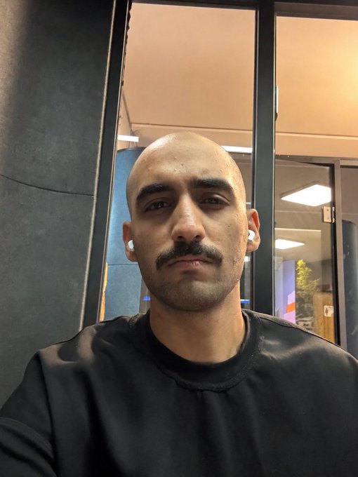 The image features a bald man wearing a black shirt with a mustache and no facial hair. He has a serious expression as he looks directly at the camera, possibly posing for a photo or video. The caption of the image includes some text that reads "just a simple smm marketer guy here, what even is zec? (all of these tickers have been ruining my life since 2019)". This suggests that the man might be a marketer or someone who has experienced negative consequences due to certain stock tickers.