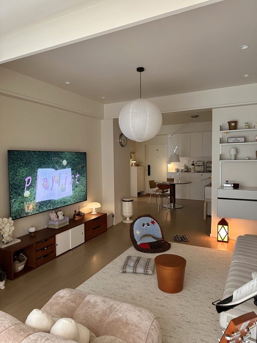 A cozy living room with a TV displaying 'playlist' on a floral background, a large paper lantern, a fluffy chair, and a view into a modern kitchen and dining area.